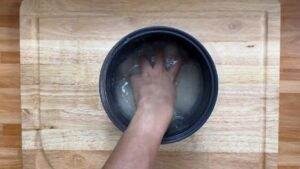 Hand rinsing rice in a pot on wooden counter, showing preparation step for a recipe.