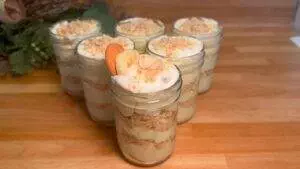 Jars of layered banana pudding with whipped cream and cookies on a wooden table.