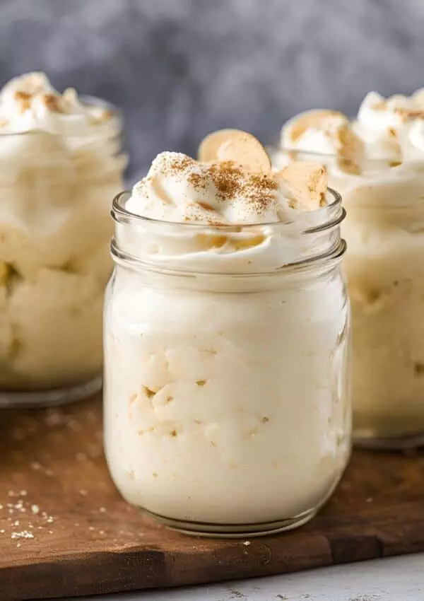 Creamy banana pudding in jars topped with whipped cream and vanilla wafers on a wooden board.