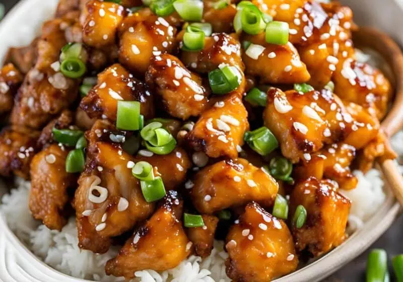 Delicious honey garlic chicken topped with green onions and sesame seeds over steamed rice in a white bowl.