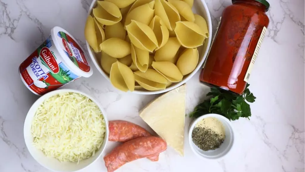 Ingredients for stuffed pasta shells: ricotta, mozzarella, sausage, pasta shells, marinara, Parmesan, herbs on marble.