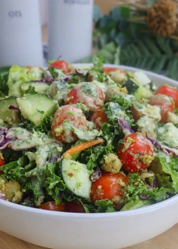 Fresh garden salad with cherry tomatoes, cucumbers, kale, and creamy dressing in a white bowl on a wooden table.