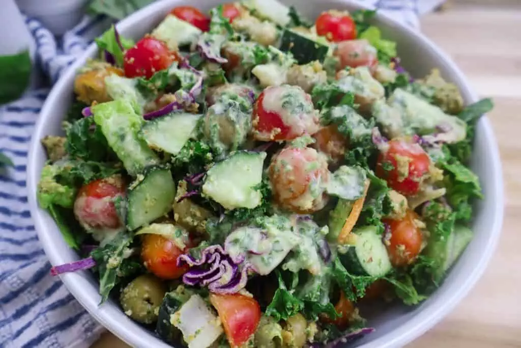 Fresh veggie salad with tomatoes, cucumbers, kale, and creamy dressing in a white bowl, perfect for a healthy meal.