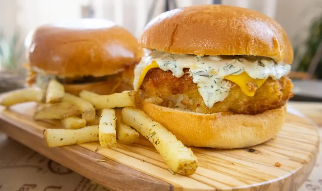 Golden crispy fish burger with cheese and dill sauce, served with seasoned fries on a wooden platter.