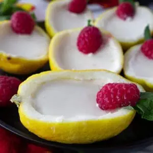 Lemon cups filled with creamy dessert, topped with fresh raspberries and mint leaves, served on a black plate.