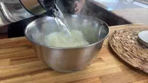 Pouring boiling water over rice noodles in a metal bowl for cooking preparation on a wooden kitchen counter.