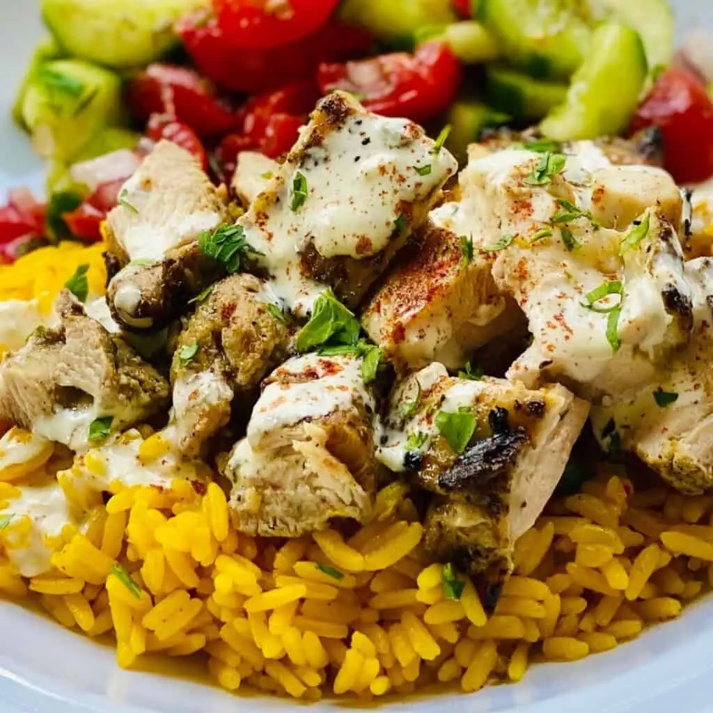 Grilled chicken with spices and herbs on yellow rice, topped with creamy sauce, served with fresh cucumber and tomato salad.