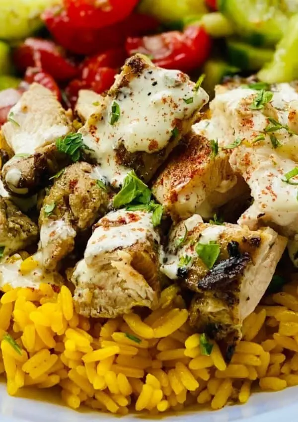 Grilled chicken with spices and herbs on yellow rice, topped with creamy sauce, served with fresh cucumber and tomato salad.