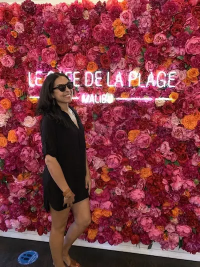 Woman in black dress posing by vibrant floral wall with Le Café de la Plage Malibu neon sign.