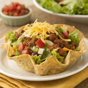 Taco salad in a crispy shell with lettuce, ground beef, tomatoes, onions, and cheese, garnished on a white plate.