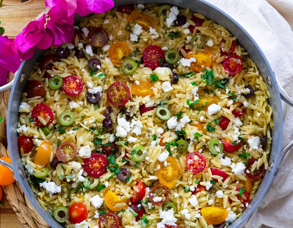 Colorful Mediterranean orzo salad with cherry tomatoes, olives, feta cheese, and fresh herbs in a skillet.