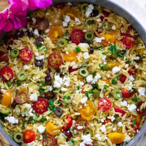 Colorful Mediterranean orzo salad with cherry tomatoes, olives, feta cheese, and fresh herbs in a skillet.