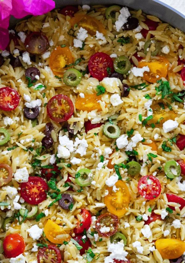 Colorful orzo salad with cherry tomatoes, olives, feta, and fresh herbs in a bowl, garnished with pink flowers.